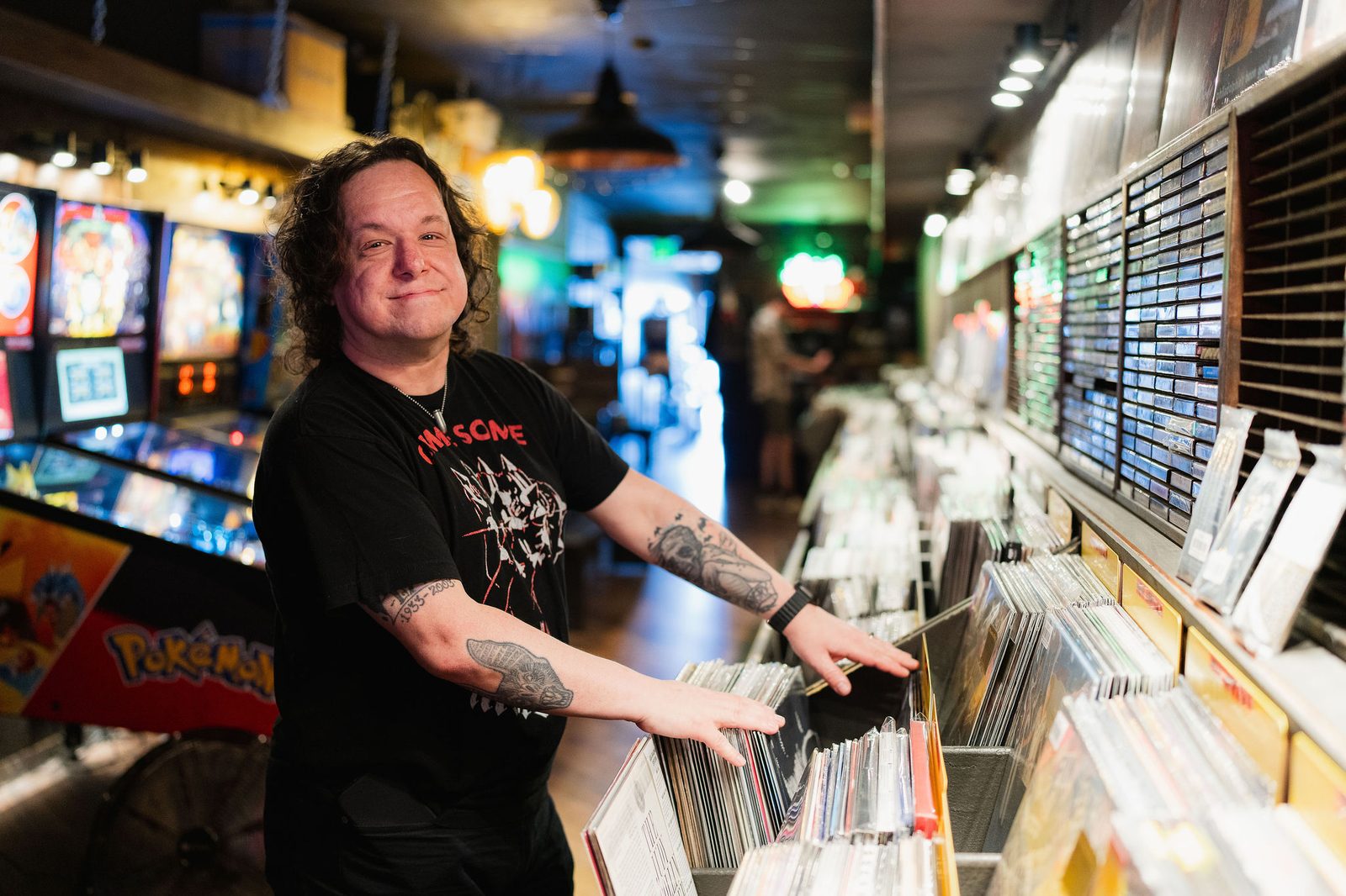 Sotirios Rebelos smiling at the record bins inside Apollo Exos Records, pinball machines glowing behind him